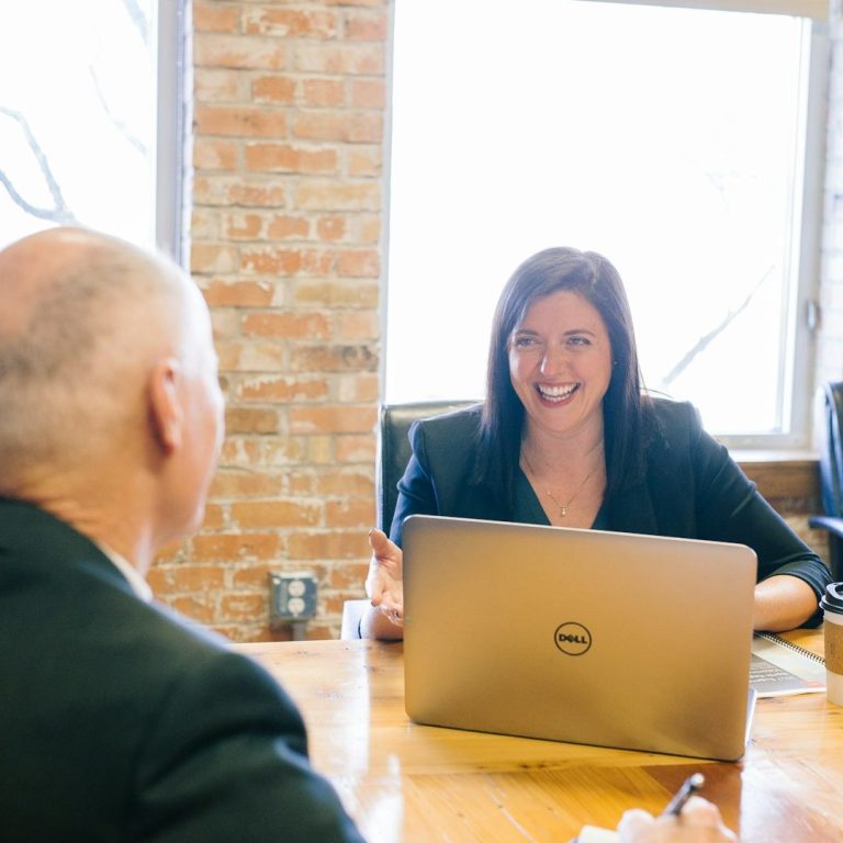 Frau im Gespräch mit einem Mann, Laptop auf dem Tisch, Backsteinwand im Hintergrund.